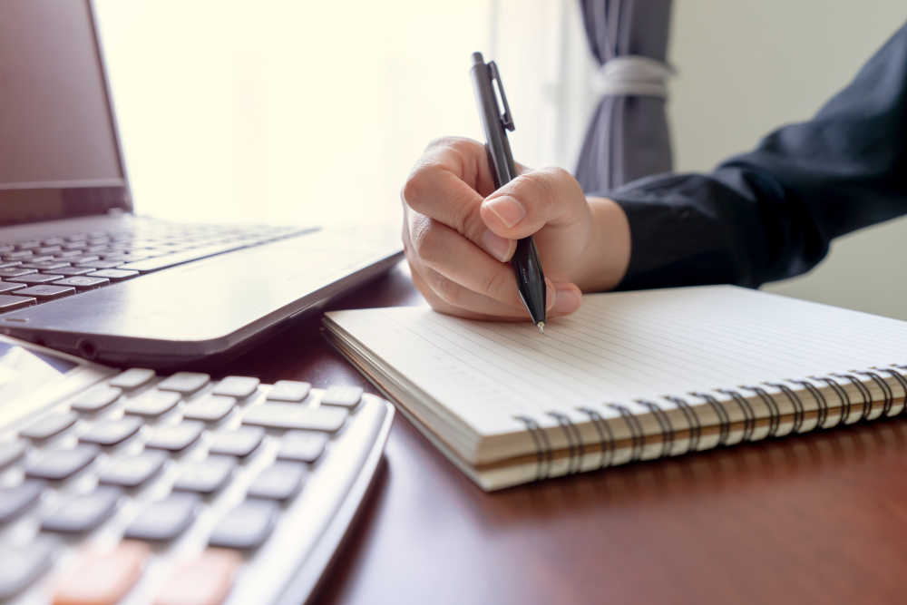 Business,Woman,Writing,Notes,In,Notebook,From,Laptop,On,Wood
